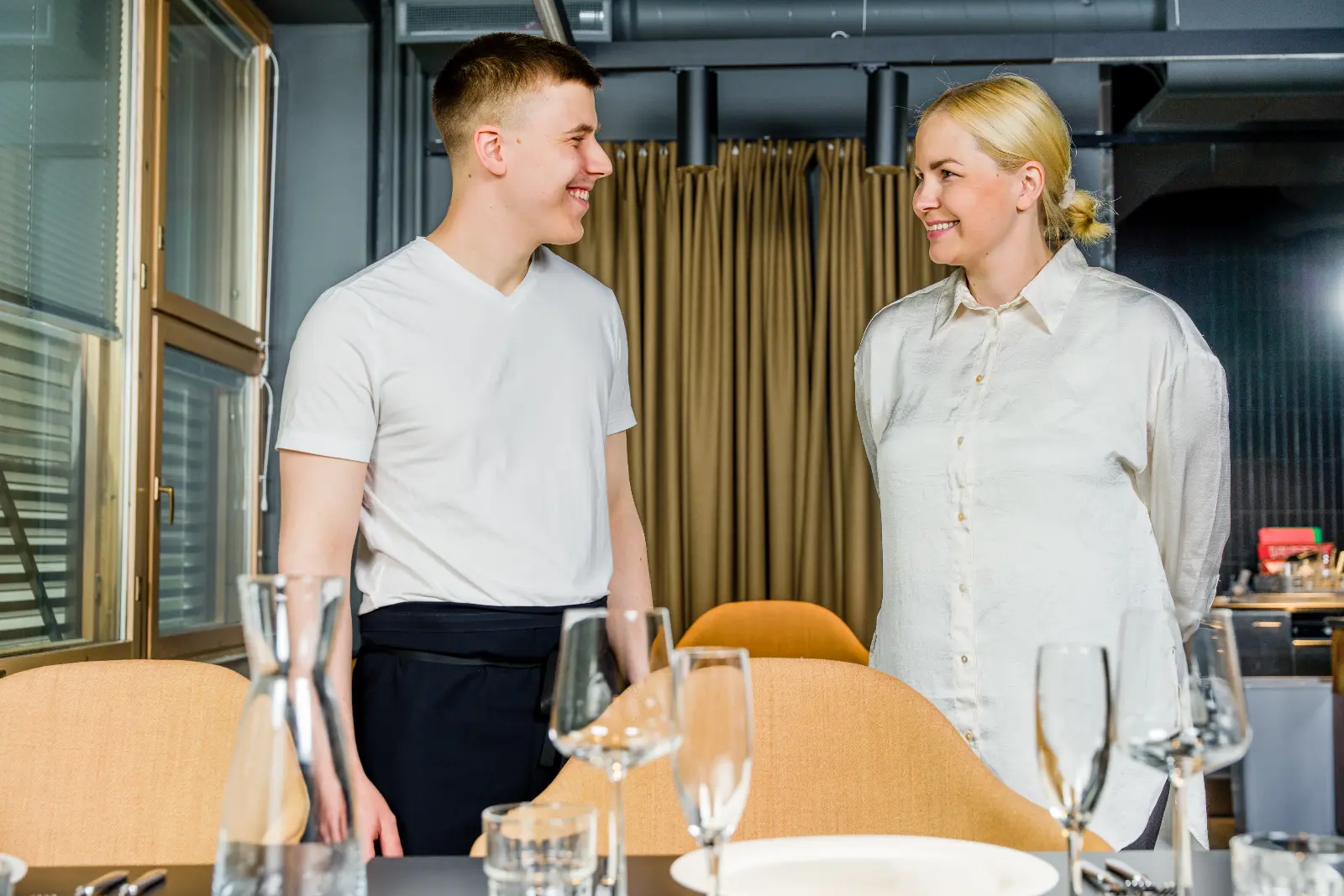 Two people in a restaurant, dressed in white shirts, looking at each other and smiling. A set table with wine glasses is in front of them.